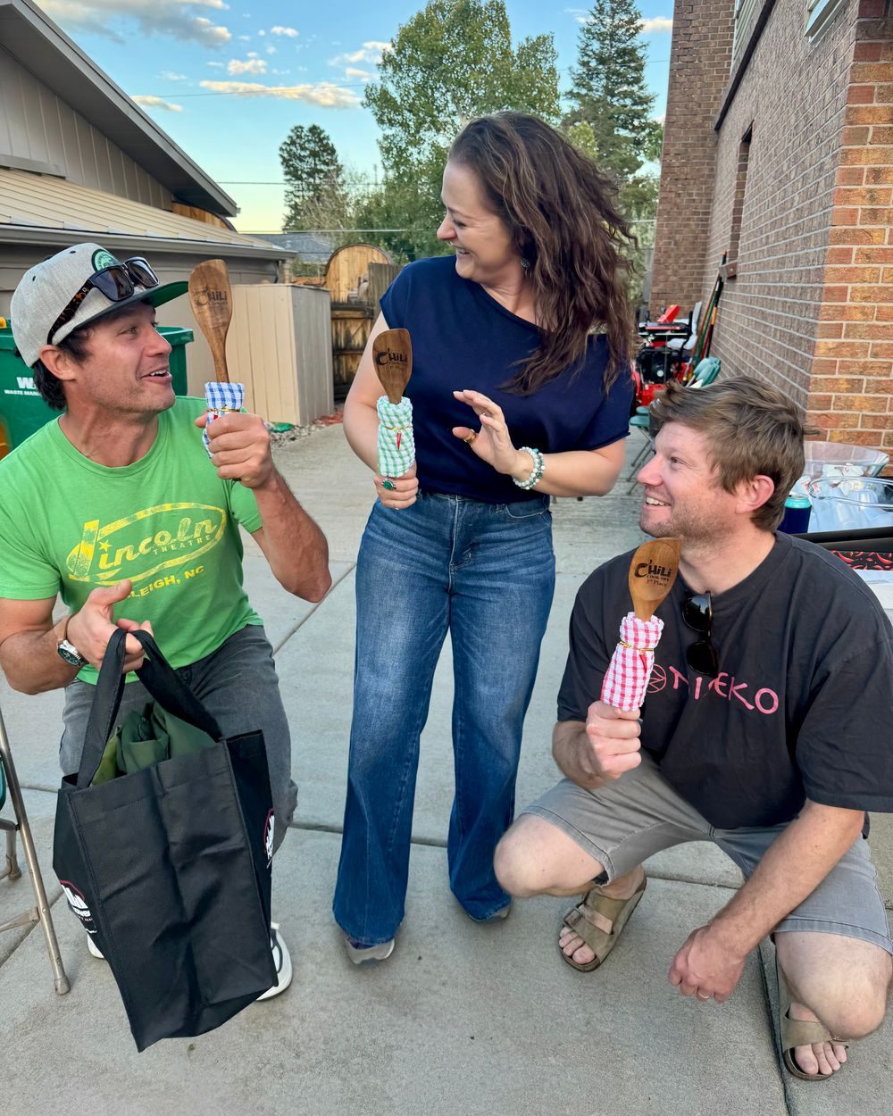Three smiling guests in a driveway holding wooden spoons wrapped in checkered cloth