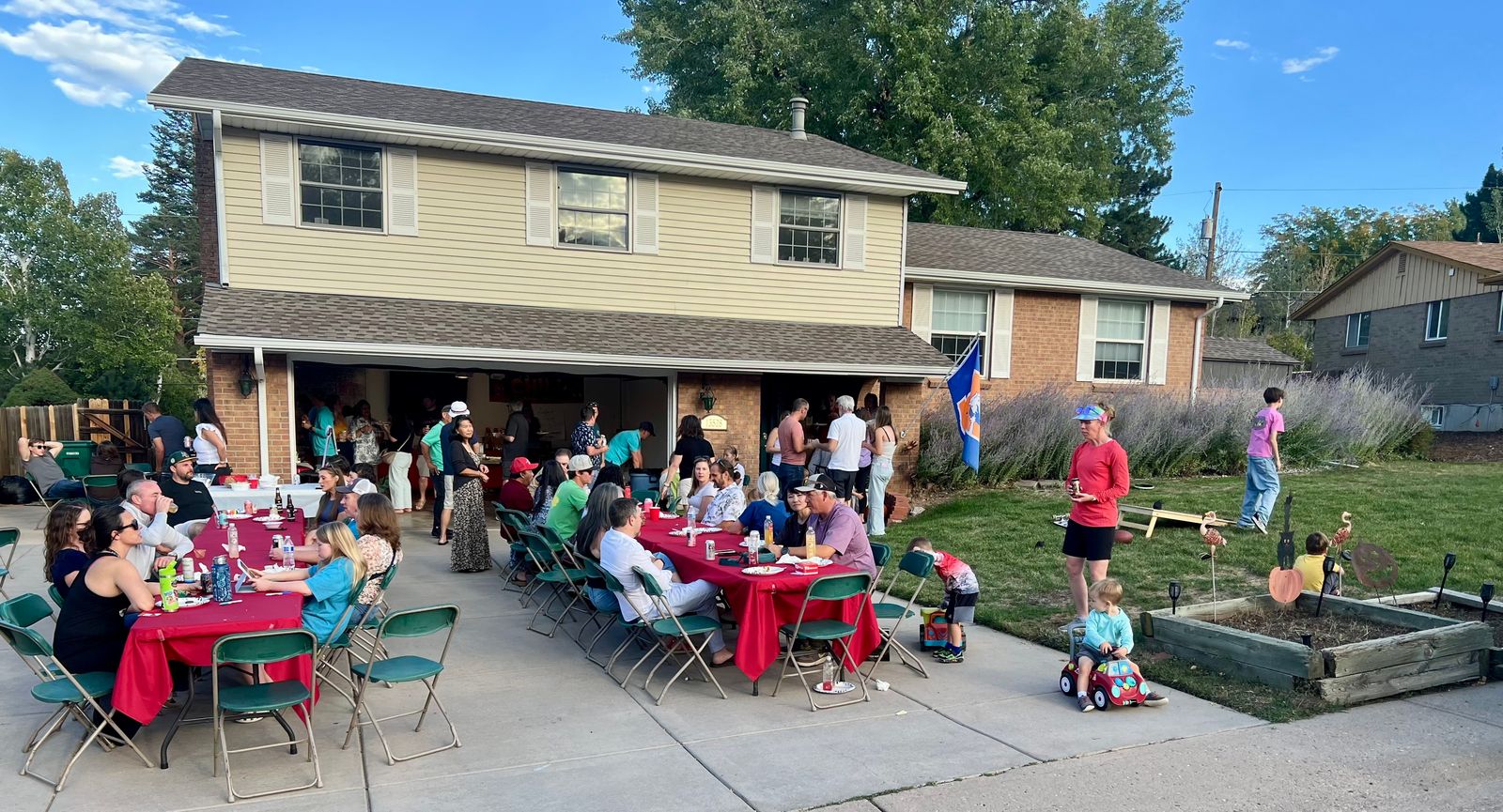 Guests at red-covered tables in a driveway in front of a two-story house, with more people in the open garage