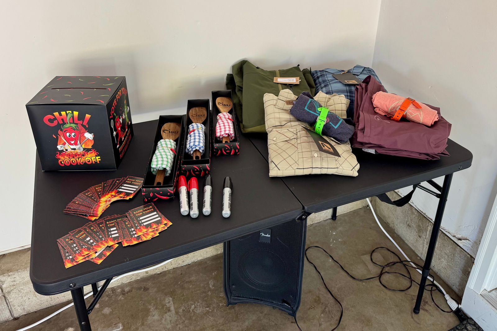 Folding table with Flylow shirts and flannels, three wooden spoons, ballot cards, markers, and the chili cook-off voting box