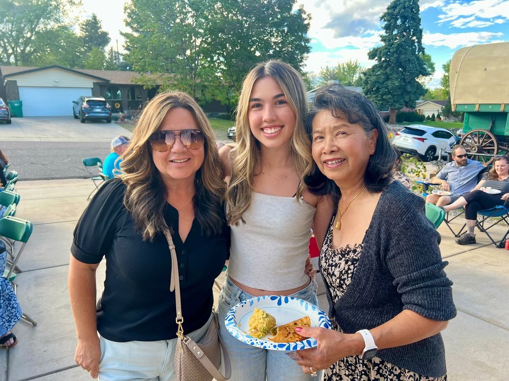 Three women smiling together outdoors, one holding a plate of chili and cornbread